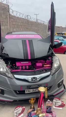 The video pans across a modified grey Honda Fit car with the hood and driver's side door open. The car features pink racing stripes on the hood and various pink accents on the engine. A banner on the windshield reads "LA PATRONA". In front of the car are several trophies and awards, suggesting it has participated in a car show. The background shows a brick wall topped with barbed wire, and other cars parked in what appears to be an outdoor lot. The overall impression is that of a custom car being displayed at an event.