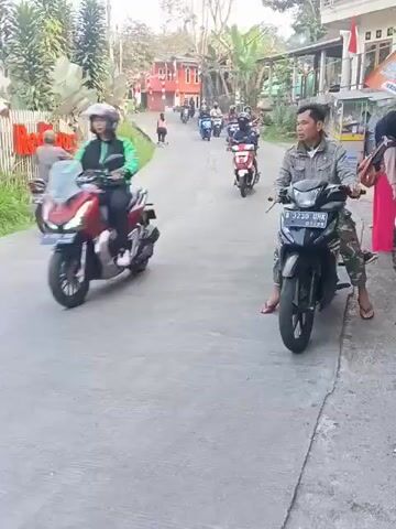 A man is riding a black motorcycle down a paved road. He is wearing a gray jacket and camouflage pants. The motorcycle has a license plate that reads "B 3230 UNK". There are other motorcycles and people on the road in the background, as well as buildings and trees. The scene appears to be in a village or rural area. The video is shot from a slightly elevated angle, looking down at the road. The lighting suggests it is daytime, possibly late afternoon due to the shadows.