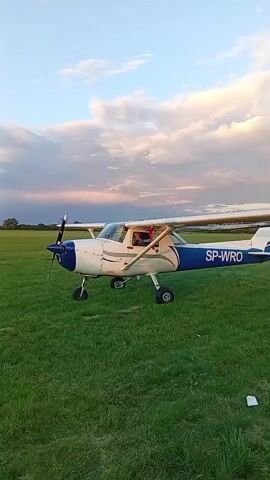 A white and blue single-engine airplane is parked on a grassy field. The airplane has the registration SP-WRO on its tail. The propeller is visible on the front of the aircraft. The sky is partly cloudy with a gradient of colors suggesting it is either sunrise or sunset. There appears to be a person in the cockpit of the airplane. The field is covered in green grass.