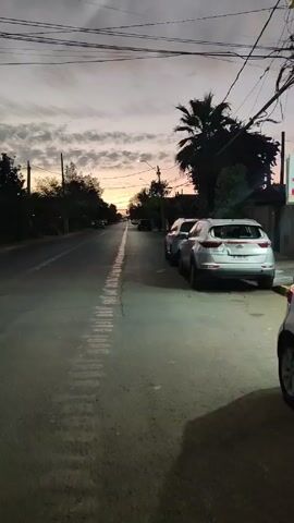 The video shows a street scene at dusk. The sky is a mix of gray and orange colors, with clouds visible. The street is paved and lined with trees and parked cars. There's a white line running down the center of the street. Power lines are visible overhead. The lighting is dim, with streetlights beginning to illuminate the scene. There are a few cars in the distance.
