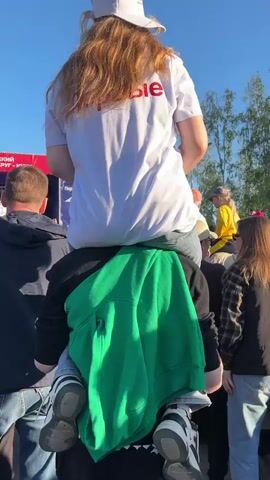 A person is carrying another person on their shoulders in a crowded outdoor area. The person on top wears a white t-shirt with red letters, light blue jeans, and a white baseball cap. Their long wavy hair is light brown. The person carrying them has a green hooded sweatshirt draped over their arms and wears dark clothing underneath. Both individuals wear sneakers. The crowd surrounding them is diverse in age and clothing styles. In the background, there is a stage with red and blue banners, a building, trees, and a clear blue sky. The scene appears to be a public event or gathering.