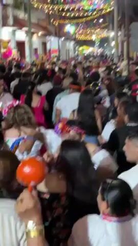 A large crowd of people are dancing and celebrating. Many of the women are wearing traditional Mexican clothing with floral headdresses. A person near the camera shakes a bright orange maraca. The event is taking place outdoors at night, under strung lights.