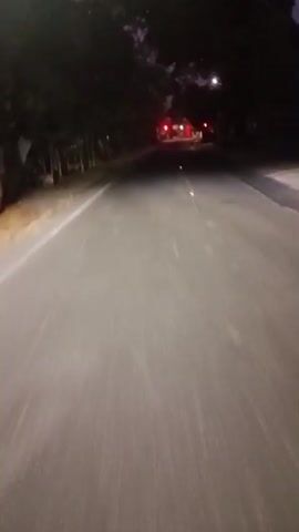 This video shows a nighttime view of a road. The road is paved and has white lane markings. There are trees and vegetation on either side of the road. In the distance, there are some lights that appear to be from buildings or streetlights. There are also red lights visible, which could be from vehicles or traffic signals. The sky is dark, indicating it is nighttime or very early morning. The camera appears to be moving, possibly from a vehicle.