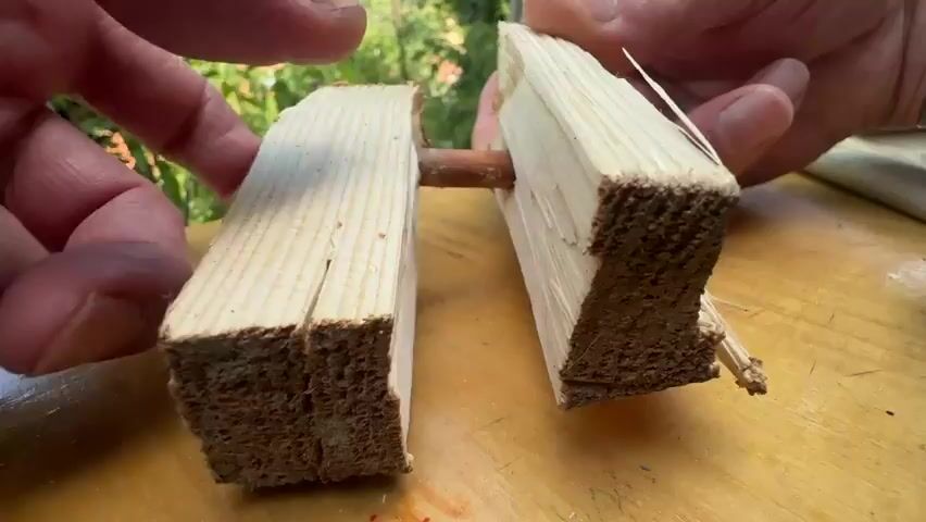 Two hands hold two pieces of rough-cut wood, each with a dowel inserted into its end. The dowels are aligned and are being pushed together. The wood appears to be pine, with visible grain and some splintering. The background is blurred but suggests an outdoor setting with greenery and sunlight.