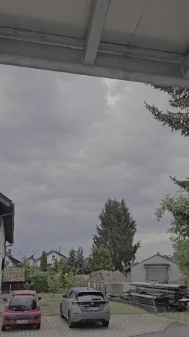 The video is an outdoor, time-lapse recording from a fixed camera perspective, looking upwards at a cloudy sky. In the foreground, the underside of a gray, metallic overhang is visible. Below the overhang, the tops of two cars are parked on a paved surface. The car on the left is red and the car on the right is gray. Behind the cars, there is a line of trees and houses with pitched roofs. A tall, dark green evergreen tree stands prominently in the middle ground. Further back, there is a building with a large, gray garage door. The sky is filled with heavy, gray clouds that appear to be moving and changing shape throughout the recording. The overall lighting is dim, consistent with an overcast day.