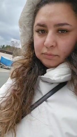 The video features a person with brown curly hair, light skin, and brown eyes wearing a white puffy jacket with a hood, standing outside on what appears to be a cloudy day. The person is looking directly at the camera, then looks off to the side, and then again directly at the camera. In the background, there is a red trailer and some greenery. The person is wearing a black strap across their chest, presumably for a bag.