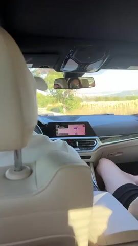 The video shows the interior of a car from a rear-facing perspective. Two people are visible in the front seats. The driver, wearing a white cap and polo shirt, is operating the vehicle. To the right of the driver, a passenger with blonde hair, sunglasses, and a black top is seated. The passenger is seen moving their arms and hands, and appears to be holding an object. Another person's legs are visible in the back seat on the right. The car is on a road with trees and vegetation on either side. The dashboard displays a navigation screen. The overall scene depicts a car journey.
