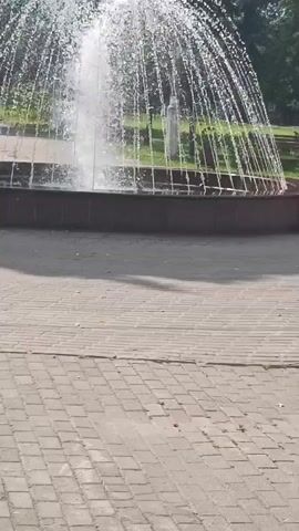 The video shows a fountain in a park. In the background, there are trees and a grassy area. There are also a few people visible in the video. One person, a young boy wearing a blue shirt and red shorts, is standing to the right of the fountain. Another person, a woman wearing a blue shirt and red shorts, is standing next to the boy. A yellow sign is also visible in the background. The fountain is spraying water into the air. The ground in front of the fountain is paved with bricks. The overall scene appears to be a sunny day in a park.