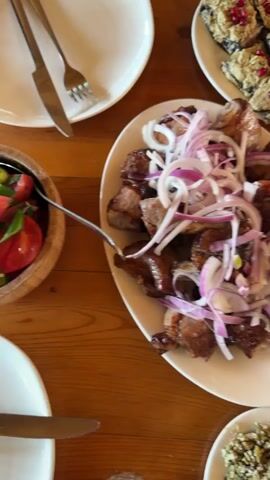 The video shows a table laden with various dishes, seemingly a meal in progress. In the center is a large white plate filled with grilled meat, topped with sliced red onions. To the upper left is a plate with eggplant slices covered in a topping and scattered with pomegranate seeds. To the upper right is a plate with slices of light-colored cheese garnished with green herbs. A wooden bowl filled with a mixed salad of chopped vegetables, including sliced jalapeños, sits near the cheese. There are also several empty white plates, silverware, and a glass of water scattered across the wooden table.  The camera briefly pans over the dishes, providing a top-down view of the table setting.