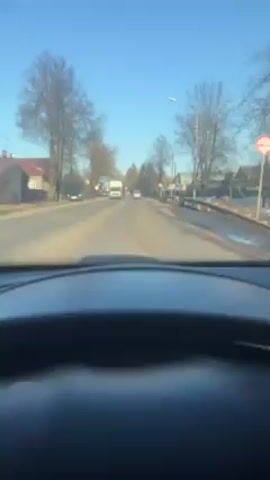 The video shows a point-of-view shot from inside a car driving down a road. The weather is clear and sunny, with a blue sky. There are bare trees on either side of the road and some houses in the background. A white truck is visible ahead on the road. The camera shows the dashboard and steering wheel of the car. A side mirror reflects the view behind the car. There are road signs visible.