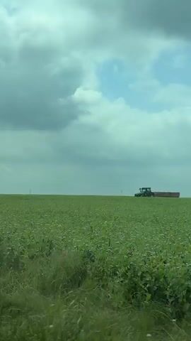 The video presents a wide shot of a vast, flat green agricultural field under an overcast sky. In the middle ground, a green tractor, resembling a John Deere, is moving from left to right across the field. It is pulling a red trailer that appears to be loaded with white material, possibly harvested crops or bales.

The scene depicts a rural, agricultural setting, characteristic of open farmlands. The field extends broadly towards the horizon. The sky is largely covered with grey and lighter blue-grey clouds, suggesting an overcast day rather than bright sunshine. The activity of the tractor and trailer indicates ongoing agricultural work, such as harvesting or transportation within the field.

The time of day is daytime. There are no people visible, and the focus is on the machinery and the natural environment. The audio includes the distinct hum of an engine, likely from the tractor, reinforcing the sense of active work. There is no visible text in the video. The location context is Izvorsko, Bulgaria.