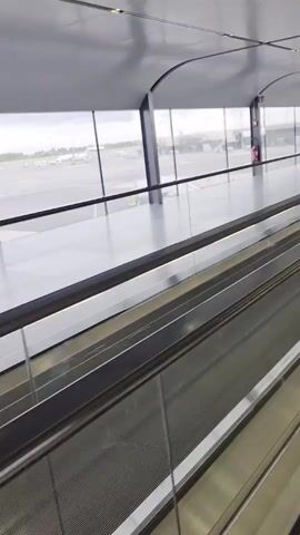 The video shows a moving walkway inside an airport terminal. The walkway is gray and metallic. It is bordered by glass panels on both sides, offering a view of the airport tarmac and aircrafts parked outside. The terminal's ceiling is gray and modern, with recessed lighting. A person in a white jumpsuit and white shoes is briefly visible standing on the walkway.