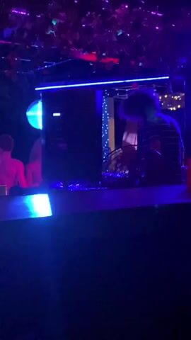 A DJ with curly hair stands behind a DJ table in a dimly lit outdoor space with blue and red lighting.  A disco ball hangs to the left of a large black speaker. A woman in red stands on the left.  Greenery hangs overhead. The DJ adjusts controls on their equipment.