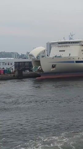 A large white and blue ferry named "Hankyu Ferry" is docked in a harbor. The ferry has multiple decks and windows. The water in front of the ferry is choppy. In the background, there are buildings and a cloudy sky. The video appears to be taken from another boat moving in the water.