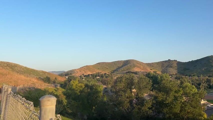 The camera slowly pans across a landscape of houses, trees, and rolling hills under a clear blue sky.  The houses are mostly light-colored with tiled roofs.  Some houses have solar panels. There is a baseball field in the distance.  The hills are green and brown with some brush. The sun is shining, creating a warm, golden light.