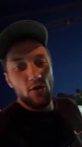 A man with a beard wearing a dark t-shirt and a baseball cap films himself and the man driving the scooter on which he is riding. The video is shot at night with street lights. He briefly turns to face the camera and sticks his tongue out while yelling.