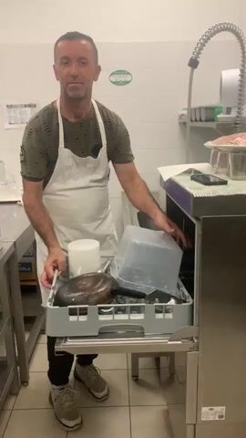A man wearing a green t-shirt and a white apron is in a kitchen. He is holding a gray plastic bin filled with kitchenware, including a large round pan and a clear plastic container. He is placing the bin onto a stainless steel cart next to a dishwasher. The kitchen appears to be a commercial establishment, with stainless steel appliances, shelving with various containers, and a commercial coffee machine visible in the background. The lighting is bright and artificial.
