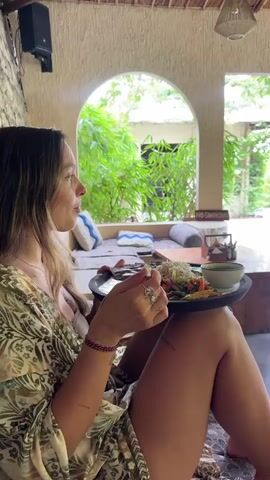 A woman sits in an outdoor lounge area, holding a plate of food in her lap and eating. She is seated on a patterned cushion, her legs visible. She is wearing a floral patterned robe and has several bracelets on her left wrist and rings on her fingers. She looks to her right. The plate contains a variety of foods, including rice, vegetables, and a small bowl of liquid. The lounge area features a stone wall behind her, a speaker mounted high on the wall, and an arched opening leading to a garden area with lush greenery. Pillows are placed on a cushioned seat in the background. A "no smoking" sign is visible. The scene appears to be set in a tropical or resort-like environment.