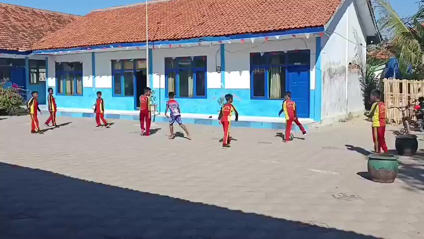 The video shows a group of young boys, dressed in red and yellow uniforms, playing soccer in the courtyard of a school. The school building has a red tile roof and blue and white walls. The courtyard is paved with gray tiles, and a flagpole stands in the center. The boys are actively engaged in the game, with some kicking the ball and others running. One boy in the foreground is focused on the ball, while another boy in the background watches them play. The scene is bright and sunny, suggesting it is daytime.