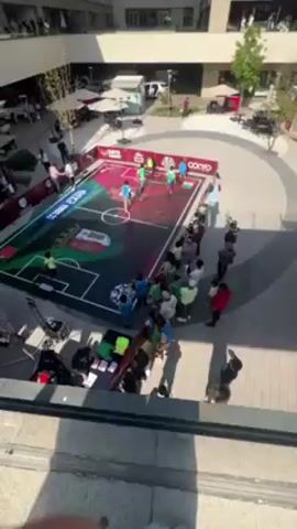 The video shows an elevated view of a street soccer event being held in an outdoor shopping mall. A small soccer field with vibrant colors and graphics is set up in the center of a circular area. Several people, presumably spectators, are standing around the perimeter of the field, watching a game in progress. There appear to be teams of players wearing blue and green jerseys playing against each other. Storefronts are visible in the background, including one that reads "Liverpool." The atmosphere seems festive and engaging.