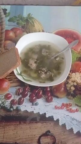 The video features a static shot of a bowl of green soup with meat and vegetables, placed on a decorative tablecloth. A slice of dark bread rests beside the bowl on the left. The tablecloth has an image of various fruits, including watermelon, apples, pears, cherries, and berries, arranged around the bowl. The surface beneath the tablecloth appears to be a wooden table with a distressed look. A spoon is visible in the soup. The lighting is bright and even, highlighting the details of the food and the tablecloth.