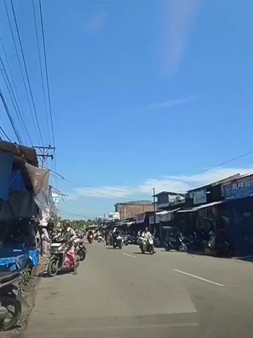The video shows a street scene with several motorcycles and a few cars. On the left side of the street, there are market stalls with people standing around. On the right side, there are buildings that appear to be shops, with motorcycles parked in front of them. The sky is clear and blue, suggesting a sunny day. Several people are visible, mostly on or near motorcycles, and some appear to be walking. The overall impression is of a busy street in what appears to be a commercial or market area.