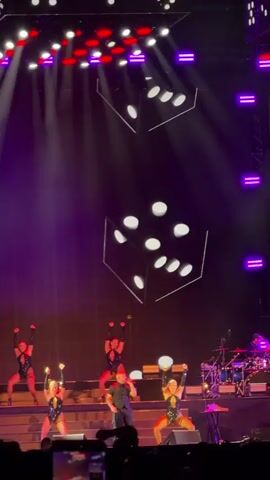 A stage performance is shown with a male singer in the center, wearing a dark shirt and pants. He is surrounded by four female dancers in black, revealing outfits with red accents. They are positioned on a multi-tiered stage, with some dancers on a lower level and others on a higher platform. Two large, projected dice are visible in the background, along with purple stage lighting and red lights on the top of the stage. A drum set and other musical equipment are visible on the far right. The audience is partially visible at the bottom of the frame. The overall atmosphere appears to be energetic, with the dancers striking poses and the singer performing.