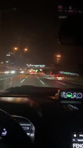 The video is filmed from the perspective of someone driving at night on a highway. The dashboard of the car is visible in the foreground, with a digital display showing various gauges and readouts. A radio is tuned to a station called "FENOMEN." Outside the car, other vehicles can be seen on the highway, their headlights and taillights illuminated. Overhead, there are streetlights and illuminated billboards. The overall impression is that of a nighttime drive through a city or suburban area.