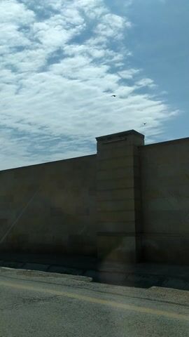 This is a video from Ghobustan, Azerbaijan, taken during the day under a partly cloudy sky. The camera is angled upwards, capturing a portion of a building with a beige stone facade. The building has an imposing, geometric design. Two decorative panels with intricate, star-shaped geometric patterns are visible on the lower section of the building, suggesting an architectural style that incorporates Islamic art. These panels are framed by stone columns and appear to be part of an entryway or facade. The surrounding area is paved, with a cracked asphalt surface and a yellow line marking a boundary, indicating it might be a road or pathway adjacent to the building. In the upper left corner of the frame, a small bird is seen in flight against the backdrop of the sky and clouds. The overall impression is of a modern, perhaps public or religious building, set against a natural sky. There is no visible text in the video.