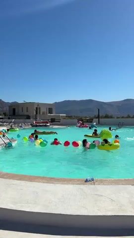 A drone shot captures a bright, sunny day at an outdoor swimming pool. The pool water is a vibrant turquoise and many people, primarily children, are enjoying various inflatable toys, including colorful balloons, rings, and animal-shaped floats like a pink flamingo and a green dinosaur. Some individuals are swimming or floating while others are sitting on the inflatables. In the background, a modern white building with multiple levels is visible, along with a mountainous landscape under a clear blue sky. The pool area has a light-colored concrete edge.