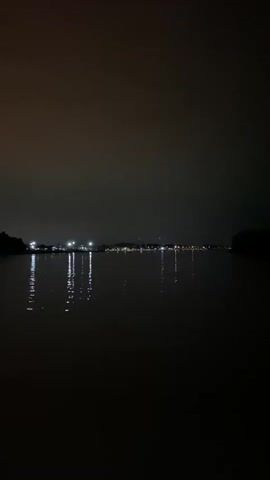 The video begins with a view of a dark body of water, reflecting city lights in the distance. The camera then moves indoors to a dark space with a crowd of people silhouetted against a red light source. Many are holding up phones, presumably recording a performance or event.