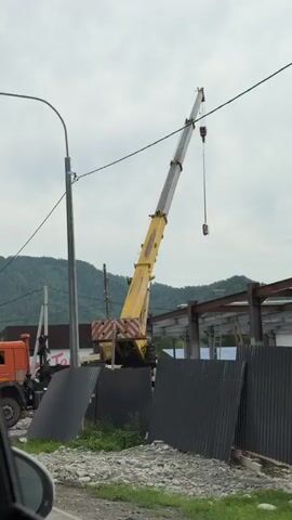 This is a vertical, medium-shot video taken from a vehicle, showing a street scene. On the left, an orange Kamaz truck is parked, and behind it, a yellow crane truck is visible. A tall street lamp is in the foreground, to the left of center. In the background, there is a building with signs in Russian. One sign reads "Март" and another, "ПРОДУКТЫ" (Products). A dark fence runs along the front of the building, and some metal sheeting leans against it. Several cars are parked on the street, including a black SUV and a white SUV with its back door open, and a man standing near it. The sky is overcast, and mountains are visible in the distance. The ground is covered in gravel.
