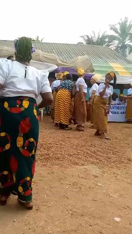 The video shows a group of women dancing outdoors, likely at an event or celebration. The dancers are dressed in colorful traditional attire, with matching headscarves and patterned skirts. They are moving in a synchronized manner, accompanied by music and singing, possibly from a nearby drum. A person in a blue and yellow spotted shirt holds a smartphone, possibly recording the performance. In the background, there is a tent with a sign that reads "ST ANTHONY OF DIEZEIKE, AMANO,". The ground is covered in reddish-brown earth and there are some buildings and palm trees in the distance.