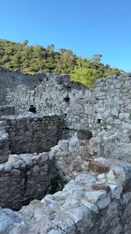 The video shows ancient stone ruins under a clear blue sky, with green, tree-covered hills in the background. The camera pans from right to left, revealing more of the archaeological site. The ruins consist of stone walls and foundations, some still standing several feet high, forming what appear to be outlines of past structures. In the middle ground, two people are seen walking towards a large, well-preserved stone doorway or archway. Other people can be briefly seen further back among the ruins. The ground is dirt and gravel. The overall impression is that of an ancient city or settlement, likely in the Kumluca region of Turkey, given the provided context. No visible text is present.