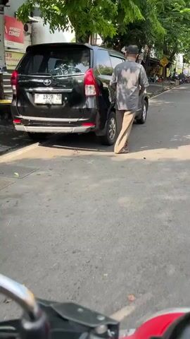 A man wearing a light-colored long-sleeved shirt and light-colored pants stands next to the rear of a black Toyota Avanza parked on the side of a street. The man has a hat on and is facing away from the camera, his right hand appears to be near the rear passenger door. The car has a license plate that reads "B 2359 ZD". A tree with green foliage overhangs the car from the left. The street is paved with asphalt, and there is a sidewalk to the left of the car. The sun is shining, casting shadows. In the foreground, a red object, possibly part of a vehicle, is visible.