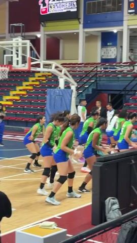 In an indoor sports arena in Ciudad Guayana, Venezuela, a group of women wearing green tops, blue shorts, black knee pads, and athletic shoes performs a synchronized dance routine on a basketball court. They move energetically, clapping and stepping in unison. Several men, some appearing to be coaches or officials, are seen observing or standing nearby.

The background features red and yellow bleachers, mostly empty. A basketball hoop and backboard are visible on the left side of the frame. Above the bleachers, a large digital scoreboard or screen displays "SANCHEZ & SALAZAR" and "TORRES" with graphic logos, along with "tantos 0" (points 0) and "jugador 0-00" (player 0-00), suggesting the event is in a pre-game or warm-up phase. Some players' jerseys have text like "A. TORRES 13" and "KOVA 6". The court features standard basketball markings, and the venue is brightly lit.
