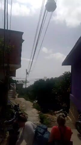 The video shows a narrow, unpaved alleyway between two buildings. The building on the left appears to be made of brick and has a balcony. The building on the right is painted purple. There are several motorcycles parked along the sides of the alley. In the foreground, two women are visible. One woman, wearing a light-colored top, is closer to the camera and appears to be looking down. The other woman, wearing an orange top, is further down the alley and is looking away from the camera. Overhead, there are several electrical wires and a large, spherical object suspended from them. In the background, there is dense foliage and a body of water, with some buildings visible in the distance. The sky is overcast.