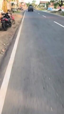 A first-person perspective video shows a paved road with a white line on the left side. Various motorcycles and a dark SUV are seen on the road, moving in the same direction. On the left side of the road, there are some market stalls, parked motorcycles, and a bicycle. The sky is clear and the lighting suggests it is either early morning or late afternoon.