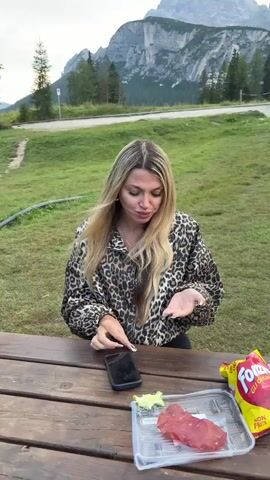 A blonde woman in a leopard-print jacket sits at a wooden picnic table outdoors. She holds a small object in her left hand and appears to be examining it. In front of her on the table are a cell phone, a clear plastic container with some raw meat and lettuce, and a bag of chips. In the background, a grassy hillside with trees leads up to a mountain range. The sky is overcast.