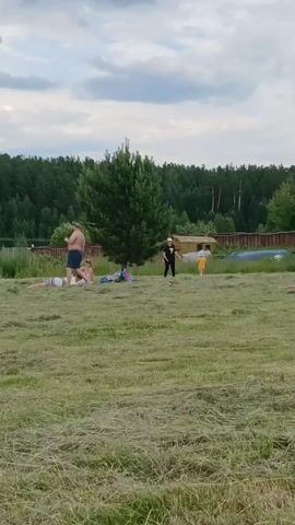 The video captures an outdoor scene on a cloudy day with a focus on a grassy field. In the foreground, freshly cut grass lies in swaths. In the middle ground, several people are visible. On the left, a man in blue shorts stands and walks towards the right. Near him, two people lie on a blanket, one wearing a white top and striped bottoms, the other in a white top and patterned bottoms. Another person is seated on a blue blanket further to the right, partially obscured by a pine tree. To the right of the tree, a person in dark clothing stands and appears to be interacting with a child in an orange outfit who is facing away from the camera. Behind them, a small wooden structure and what appears to be a boat or canoe are visible. The background consists of a dense line of trees and a cloudy sky. The overall impression is of a casual outdoor gathering or recreation.