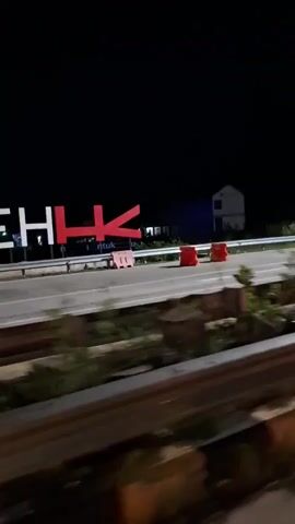 The video shows a dark night scene with a large white and red sign that reads "BANDA ACEH" in a sans-serif font. The letters "H" and "K" in "ACEH" are red. The camera is moving horizontally, revealing the full sign, which is elevated on a metal structure. Below the sign is a two-lane road with yellow and white markings. Several orange and white plastic barriers are visible on the side of the road. In the background, there are some blurry buildings and structures. The ground is dark, possibly asphalt. The overall impression is that of a roadside sign at night, possibly indicating a city entrance or landmark.