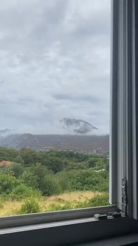 A time-lapse video shows a mountainous landscape under a cloudy sky. In the foreground, a line of green trees and bushes is visible, with a hint of yellow grass. In the middle ground, more trees and some reddish-brown vegetation cover the lower slopes of the mountains. The upper parts of the mountains are rocky and appear to be partially obscured by thick, white clouds. The sky is filled with gray, textured clouds, creating a moody atmosphere. The video captures the subtle shifts in the cloud cover and light over the landscape. The camera appears to be positioned behind a window frame, as indicated by the vertical white structure on the right side of the screen.
