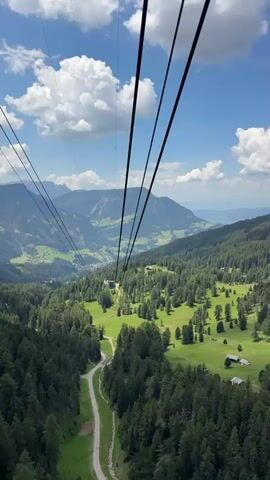 This media file captures a scenic view from a cable car ascending a mountain in Urtijëi, Italy. The primary subjects are the cable car's thick black cables, which dominate the foreground and stretch diagonally across the frame, guiding the viewer's eye towards the expansive landscape. The scene is a lush, mountainous region characterized by dense green forests covering the slopes, interspersed with open, vibrant green meadows dotted with small, traditional chalets. A winding dirt road is visible in the lower left, disappearing into the trees.

The activity taking place is the ascent via cable car, offering breathtaking panoramic views. The time of day appears to be daytime, likely late morning or early afternoon, given the bright sunlight and the shadows cast by the trees and clouds. The weather is fair, with a clear blue sky punctuated by fluffy white cumulus clouds, suggesting a pleasant and warm day. The location is clearly mountainous, with layers of rolling hills and more distant, rugged peaks visible in the background. The overall emotion evoked is one of awe and tranquility, thanks to the stunning natural beauty and the peaceful movement of the cable car. No visible text is present in the media file.