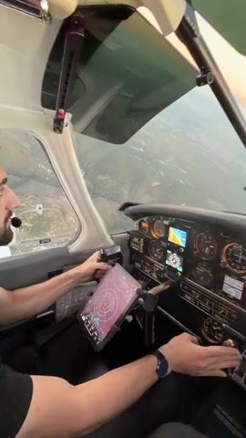 The video shows the interior of a cockpit of a small aircraft during flight. Two individuals are visible, both appearing to be pilots or passengers. One person, on the left, is seen with their hands on the yoke and a tablet mounted in front of them. The other person, on the right, has their hands near the throttle controls. The dashboard displays various instruments and screens, including navigation information on the tablet. Outside the cockpit windows, the landscape and the horizon are visible, suggesting the aircraft is in the air. The lighting and sky indicate it might be during sunrise or sunset, with a gradient of colors visible on the horizon. The camera perspective is from within the cockpit, moving slightly, giving a dynamic view of the flight.