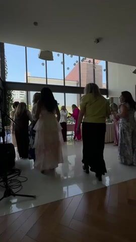 A group of women are gathered in a well-lit room with large windows. Most of the women are wearing dresses. Some are dancing, while others are standing and observing. The room appears to be decorated for an event, with some sort of string lights or decorations hanging from the ceiling. A speaker and sound equipment are visible in the foreground on the left. The floor is a polished wood laminate. The overall atmosphere appears to be festive and celebratory.
