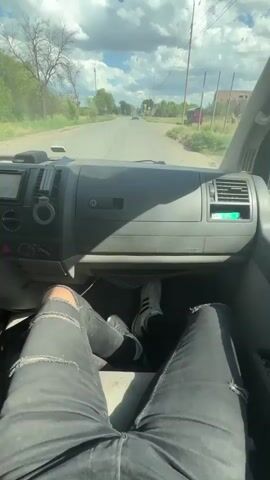 The video shows a first-person perspective from inside a vehicle, likely a van, as it drives down a paved road. The camera is positioned to show the dashboard, the road ahead, and the legs of the driver and possibly a passenger. The driver and passenger are both wearing ripped black jeans. The passenger's feet are visible on the floor, wearing white sneakers with black stripes. The dashboard has a central console with various buttons and air vents. The road is lined with trees and some houses on either side. The sky is cloudy with patches of blue. There are traffic signs visible along the road. The vehicle appears to be moving at a moderate speed.