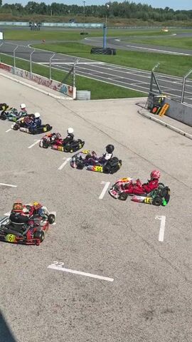 The video shows a go-kart race starting lineup. Several go-karts, each with a driver wearing a helmet and racing suit, are lined up on a paved track. The karts are of various colors and designs, and some have visible numbers. The track appears to be outdoors, with a grassy area and fencing visible in the background. The sky is clear and blue, suggesting a sunny day. The go-karts are stationary, and the drivers are seated in their karts, seemingly waiting for the start of the race. Some drivers appear to be looking forward, while others might be looking to the side. The perspective is from above and slightly to the side of the lineup, offering a clear view of the karts and drivers.