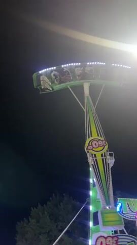 This video shows a carnival ride at night. The ride is tall and green and white, with lights. People are visible on the ride, which is swinging back and forth. The ride appears to be in motion. There are trees in the background.