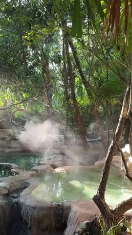 The video shows a natural hot spring nestled within a lush forest. Steam rises from the surface of the turquoise water, creating a mystical atmosphere. The hot spring is surrounded by rocks and vegetation, with sunlight filtering through the trees, illuminating the scene. The water appears to be shallow and clear, with some rocks visible beneath the surface. The surrounding forest is dense with green foliage and tall trees, creating a sense of seclusion and tranquility. The overall impression is one of a serene and secluded natural paradise.
