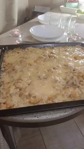 The video shows a close-up of a casserole in a dark metal baking dish on a kitchen counter. The casserole is topped with melted, golden-brown cheese, and appears to contain potatoes or other vegetables underneath. To the left of the casserole is a small, lit candle in a glass holder. Further back on the counter, there are plates, glasses, and a bowl of green apples, suggesting a meal is being prepared or served. The overall environment appears to be a home kitchen. The camera is stationary and focused on the casserole.